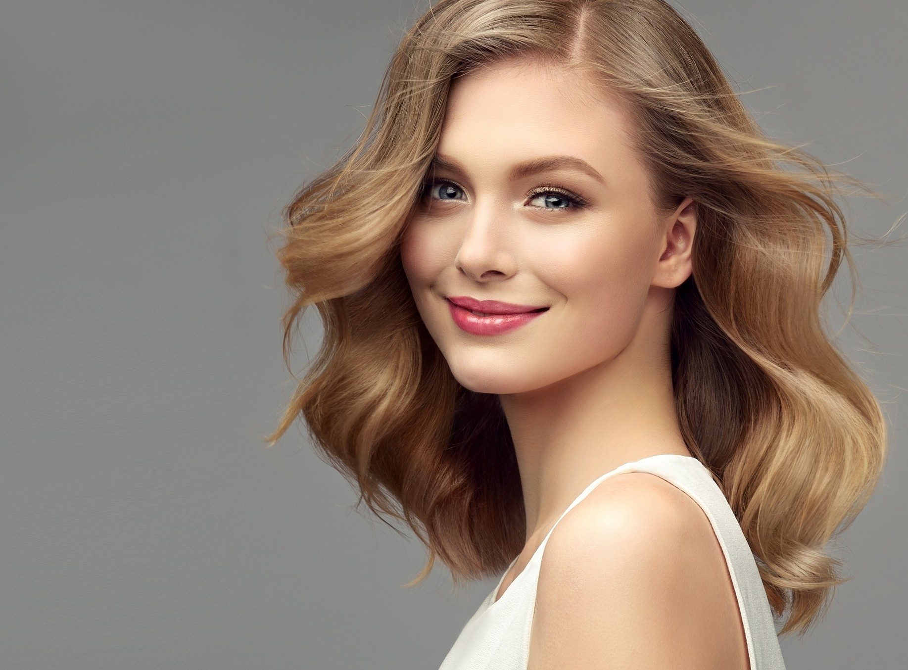 Model with dark blonde hair. Frizzy, elegant hairstyle is surrounding lovely face of tenderly smiling young woman. Natural gloss and softness of healthy hair. Hair care and hairdressing art.